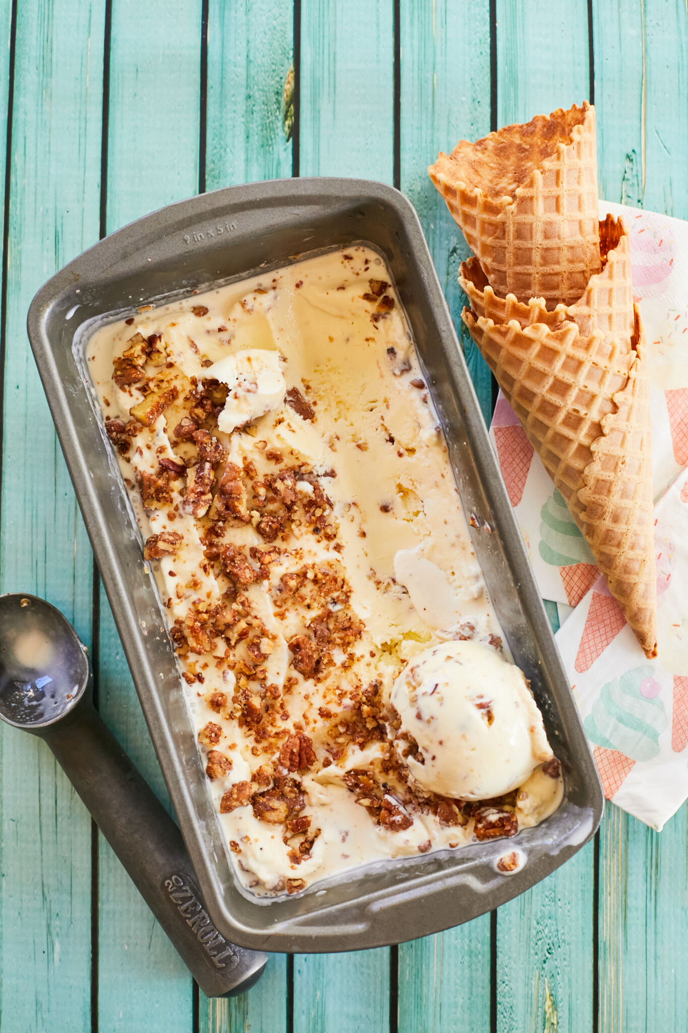 Extra smooth pecan gelato in a metal loaf pan, with crunchy toasted nuts on top and mixed through out. One ball is scooped on the right side in the pan. The scoop is on the let side and ice cream cones are on the right side outside the pan. 