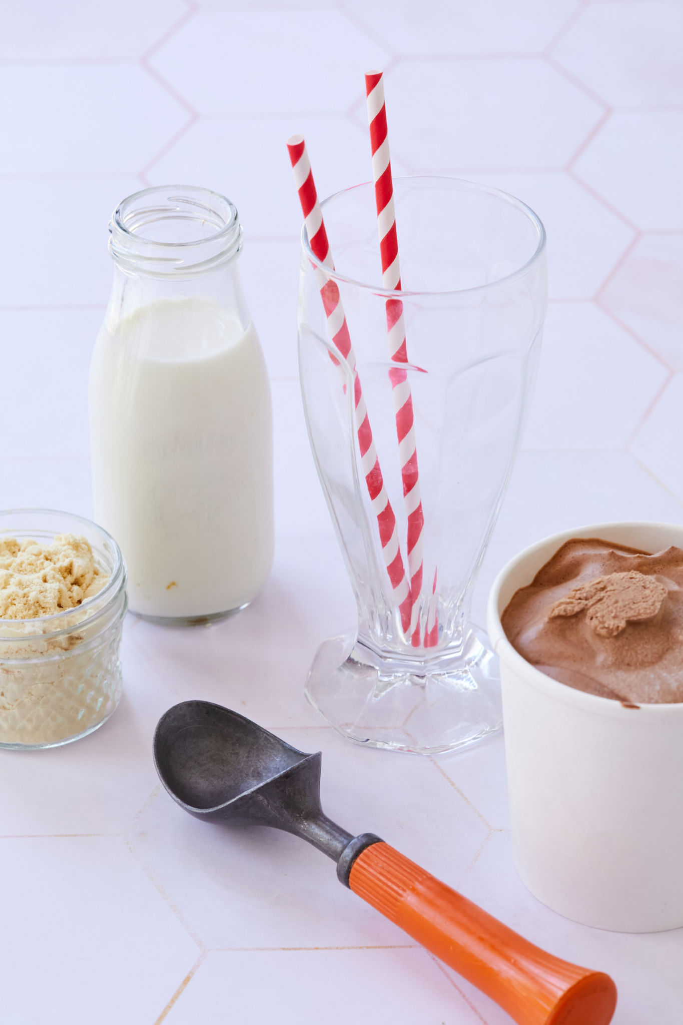 An empty soda fountain glass with two red and white straws sits next to a glass of milk, a jar of malt powder, and a tub of chocolate ice cream.