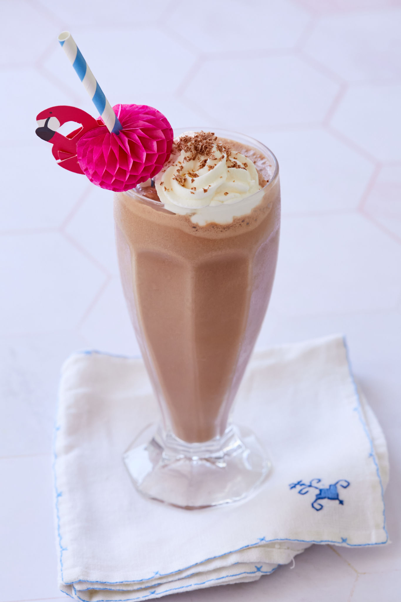 A chocolate malt milkshake is garnished with whipped cream in a tall glass with a blue and white straw and festive flamingo straw decoration.