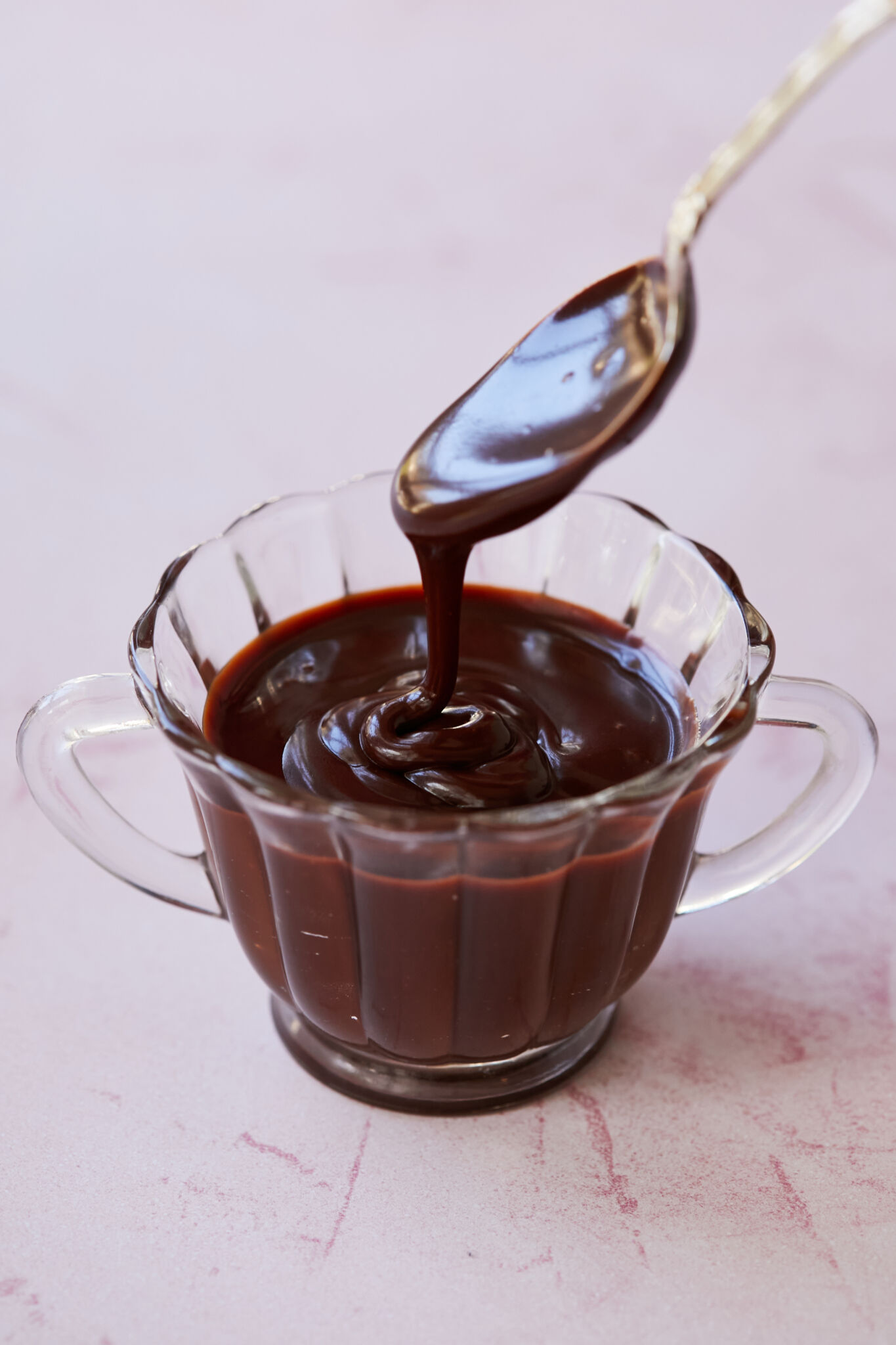 Silky smooth Chocolate caramel sauce is dripping from a spoon into a glass bowl with two handles. 