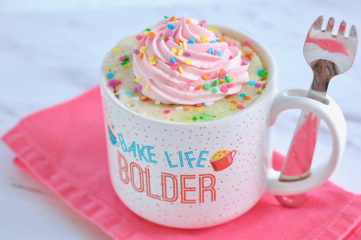 Made in under five minutes, this is my Best-Ever Vanilla Mug Cake in a Gemma's Mug Meals Mug with Spork in the handle, topped with pink frosting and sprinkles.