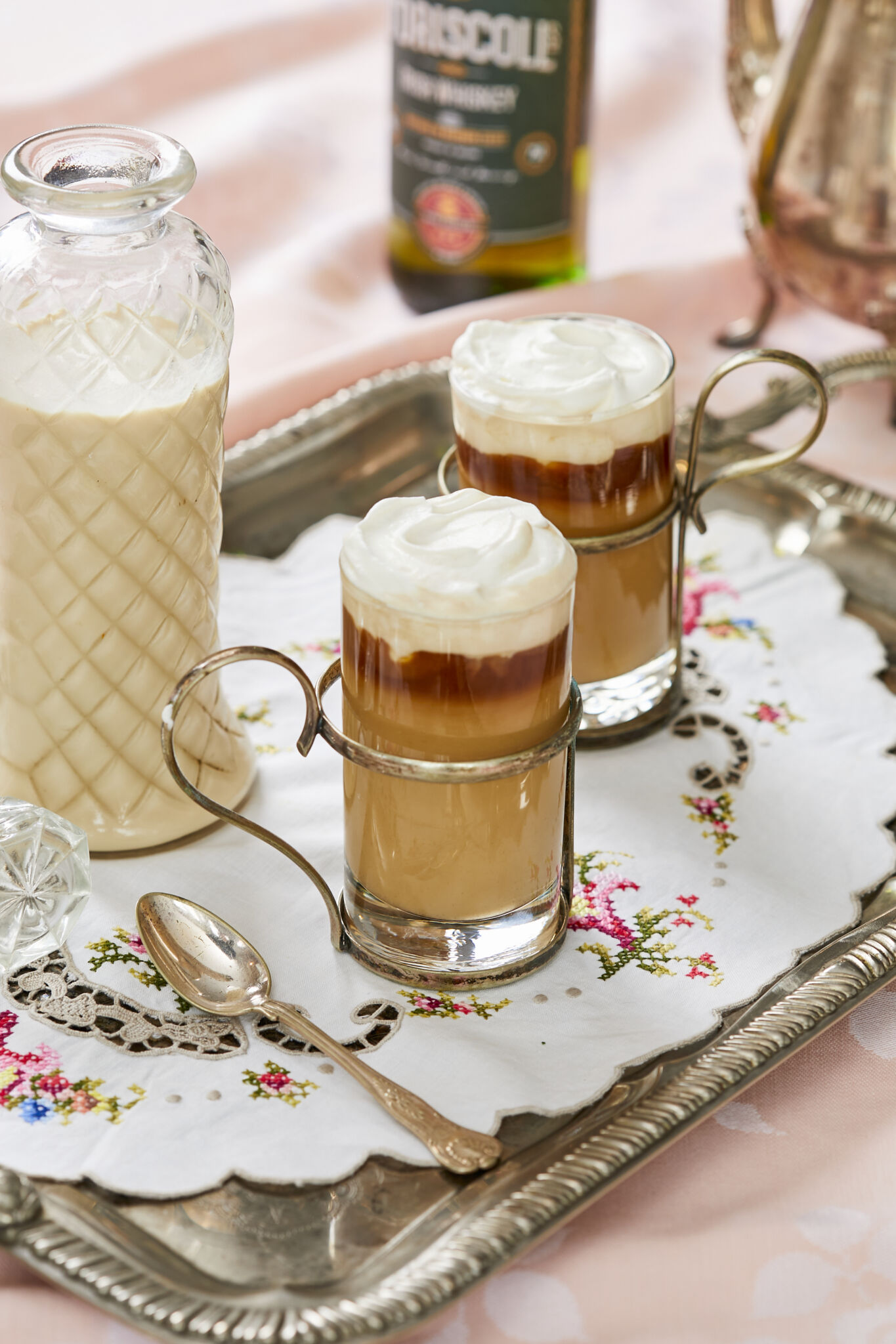 Silky smooth Homemade Irish Cream is in stored in a tall weave pattern glass bottle on the left side on a silver platter. To the right of the bottle are two glasses of Irish Cream Frappuccino which has blended sweet, floral, nutty and boozy Irish Cream with coffee and milk at the bottom, topped with pillowy whipped cream. A silver spoon is also on the platter next to the glasses. A bottle of O'Driscoll's whiskey and a tea pot are next to the platter in the upper right corner of the image. 