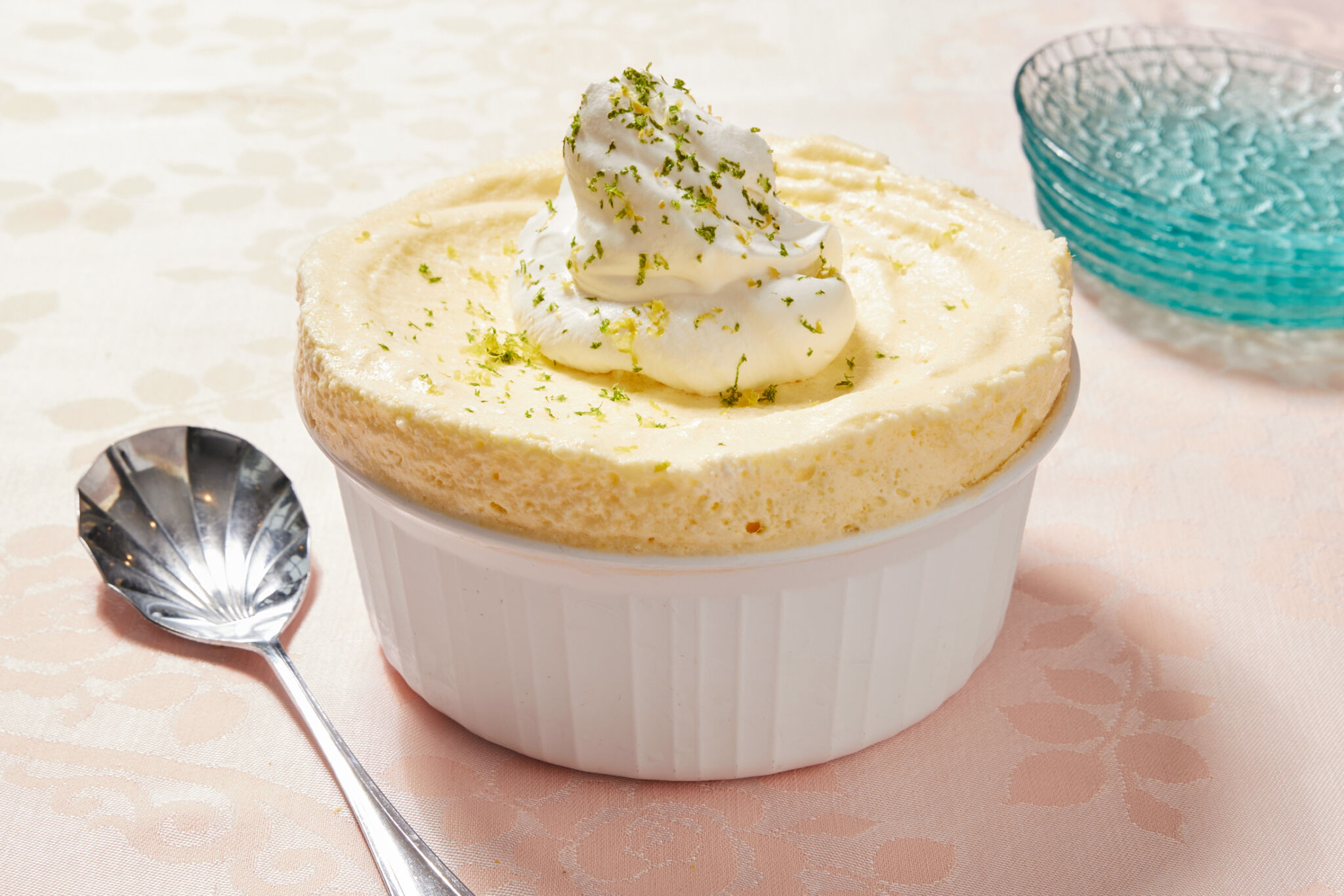 The silky smooth soufflé is perfectly set in a white ramekin, topped with a dollop of fluffy whipped cream and sprinkled with refreshing lemon and lime zest to boost the flavor. A silver serving spoon is in the bottom let corner and a stack of light-blue glass dessert plates are in the top right corner.