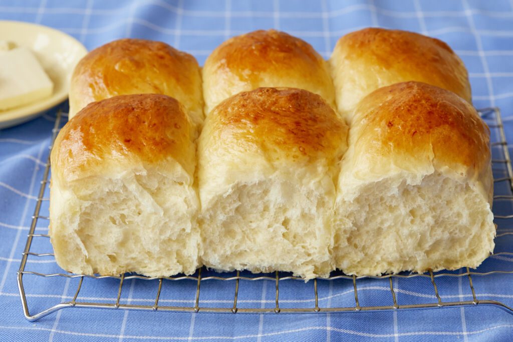 A close shot at Mashed Potato Dinner rolls which are cooling or a wire rack shows their soft and bubbly crumb and golden brown top. The rolls are served with butter on a small plate.