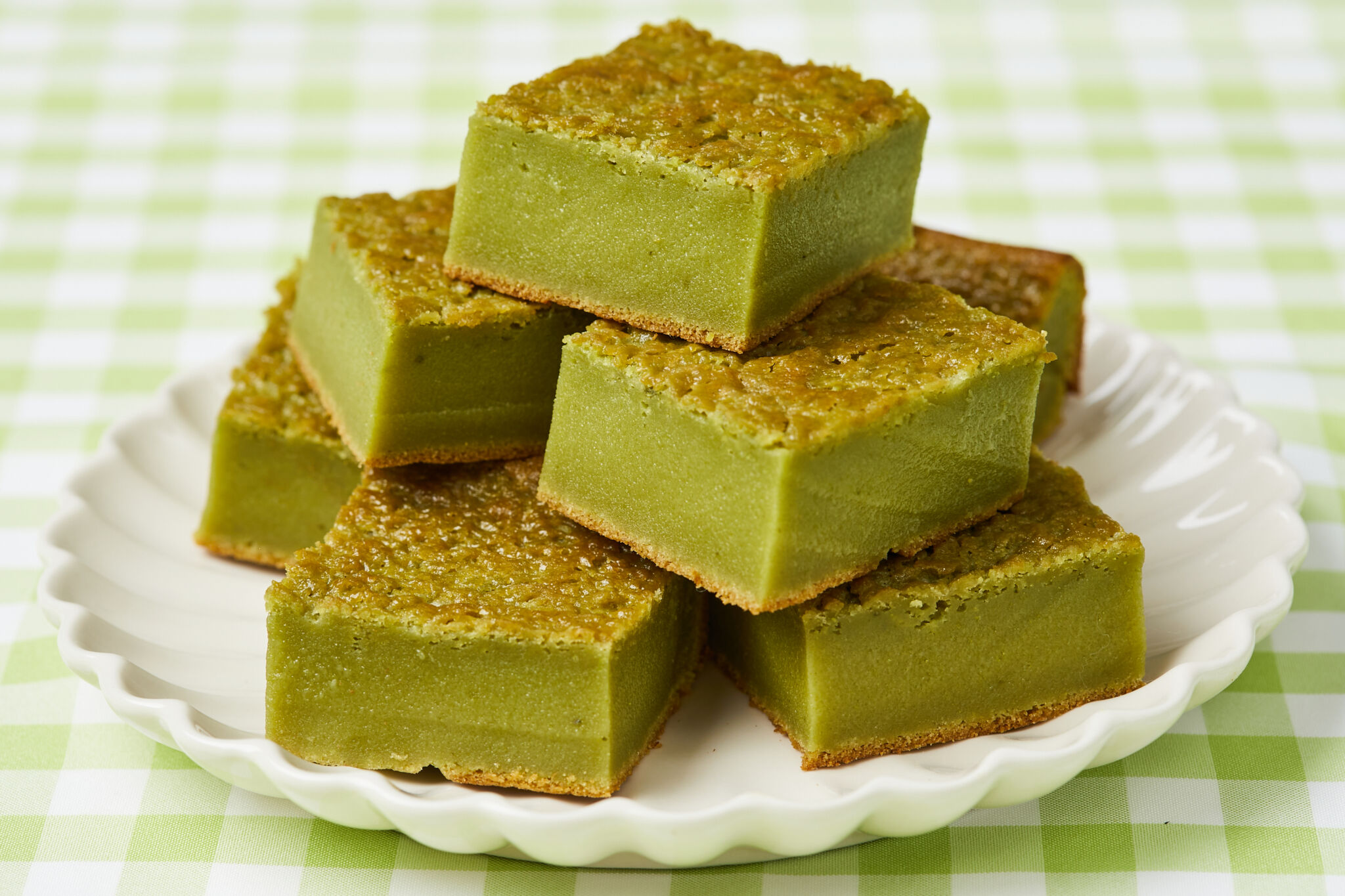 6 squares of rich Matcha Butter Mochi are placed on a white scalloped-edge plate. The green tea sweet rice cake has golden crispy crust on top and at the bottom, with green smooth chewy and soft center.