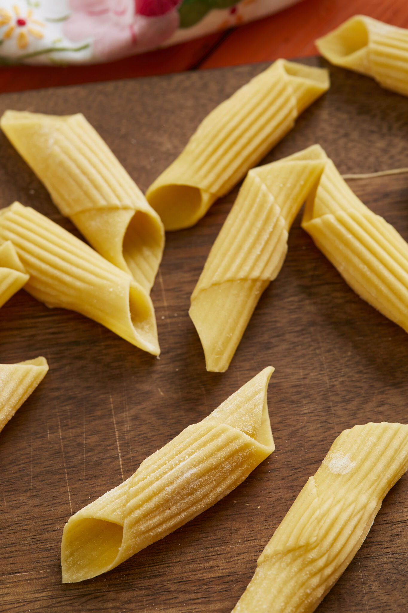 Penne pasta is drying on a wooden board. It's in a tubular-shape cut diagonally on the ends and resembles an old-fashioned writing quill that was dipped in ink. 