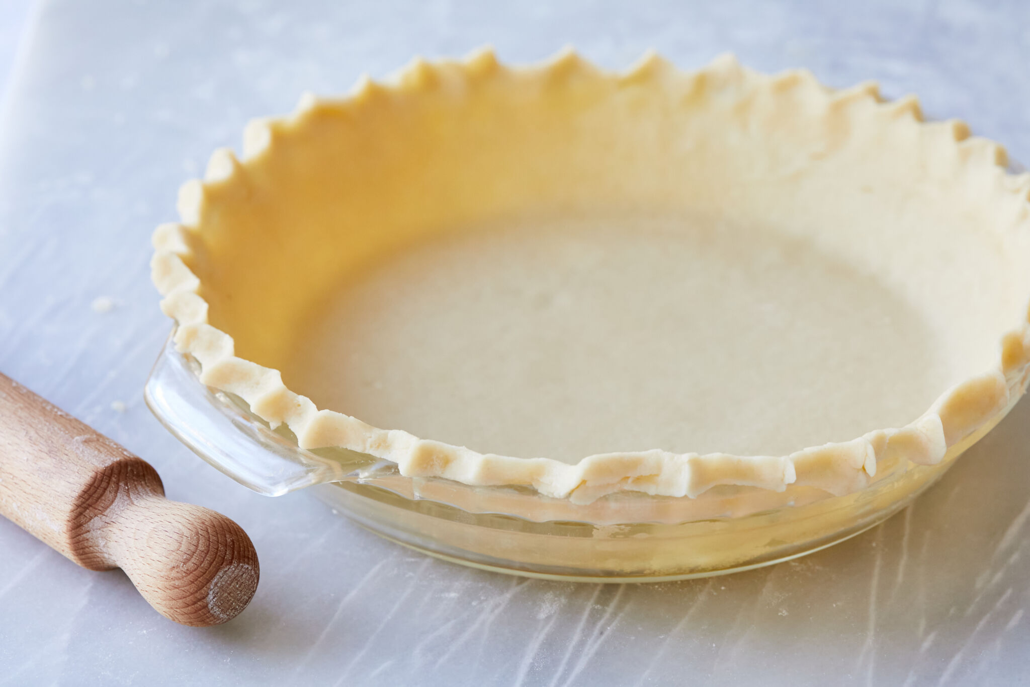 A Flaky Sour Cream Pie Crust is lined in a glass pie dish , with evenly crimped edges. A wood rolling pin is placed next to the pie crust in the left bottom corner of the image.