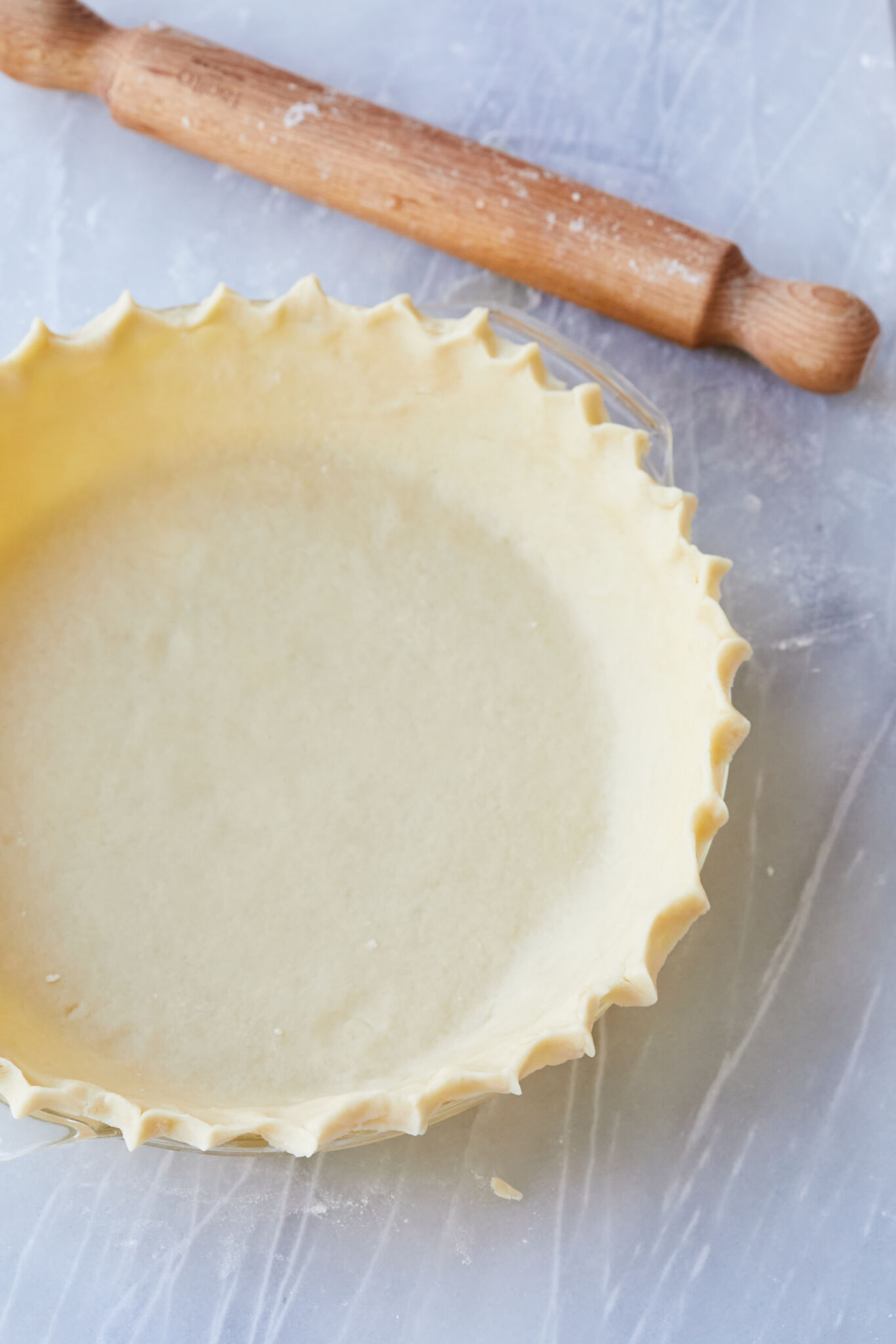 A smooth Flaky Sour Cream Pie Crust is lined in a glass pie dish , with evenly crimped edges. A wood rolling pin is placed next to the pie crust. 