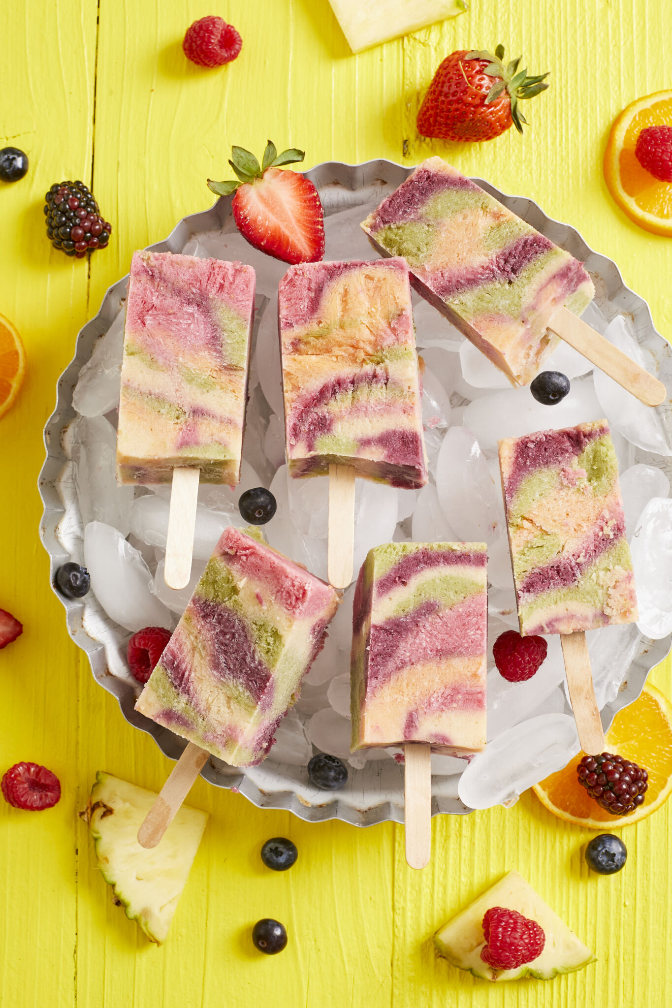 Colorful all natural tie dye popsicles are in orange, red, purple, and green colors, served on ice in an aluminum pie pan, with fresh blackberries, raspberries, strawberries, oranges, pineapple on the side.