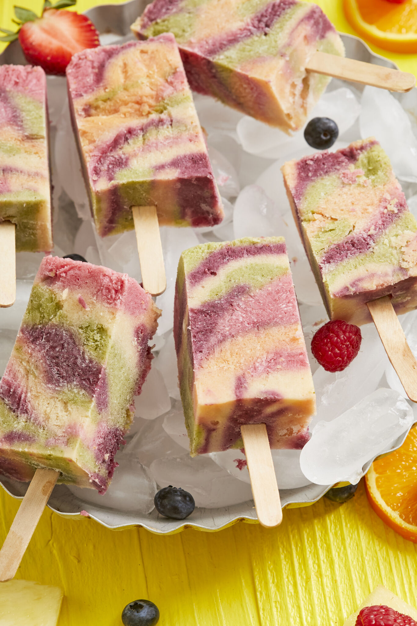 The all-natural frozen treats are in a beautiful tie-dye pattern with red, pink, yellow and green colors, served on ice cubes in a metal pie pan, with blueberries, raspberries and orange slices on the side.