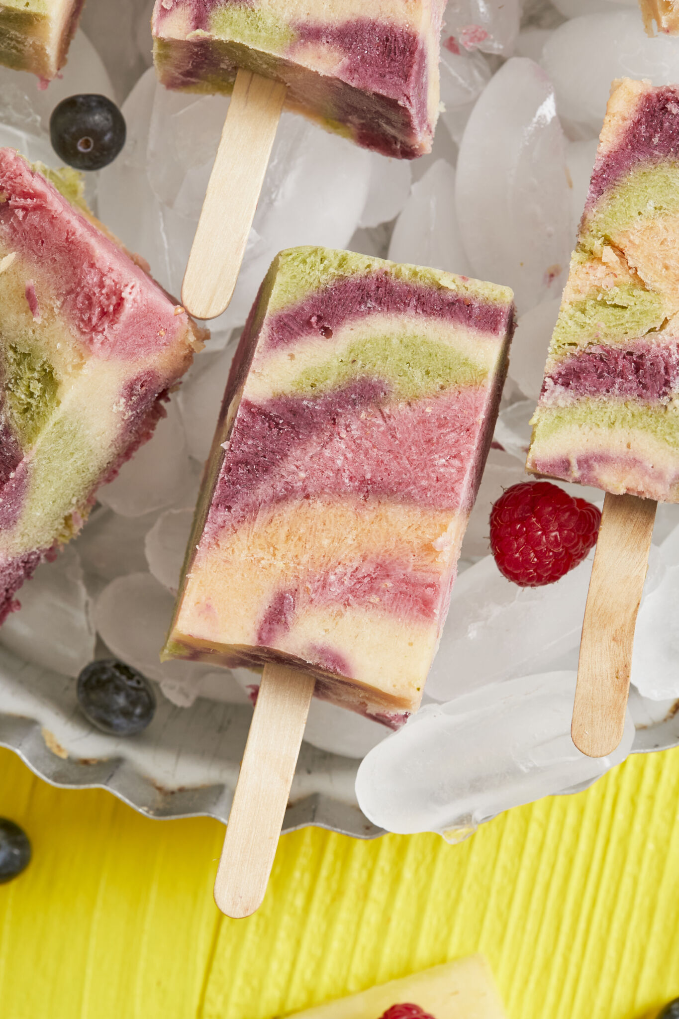 A close shot of frozen treats in beautiful trippy pattern with red, pink, yellow and green colors, served on ice cubes in a metal pie pan, with blueberries and raspberries on the side. 